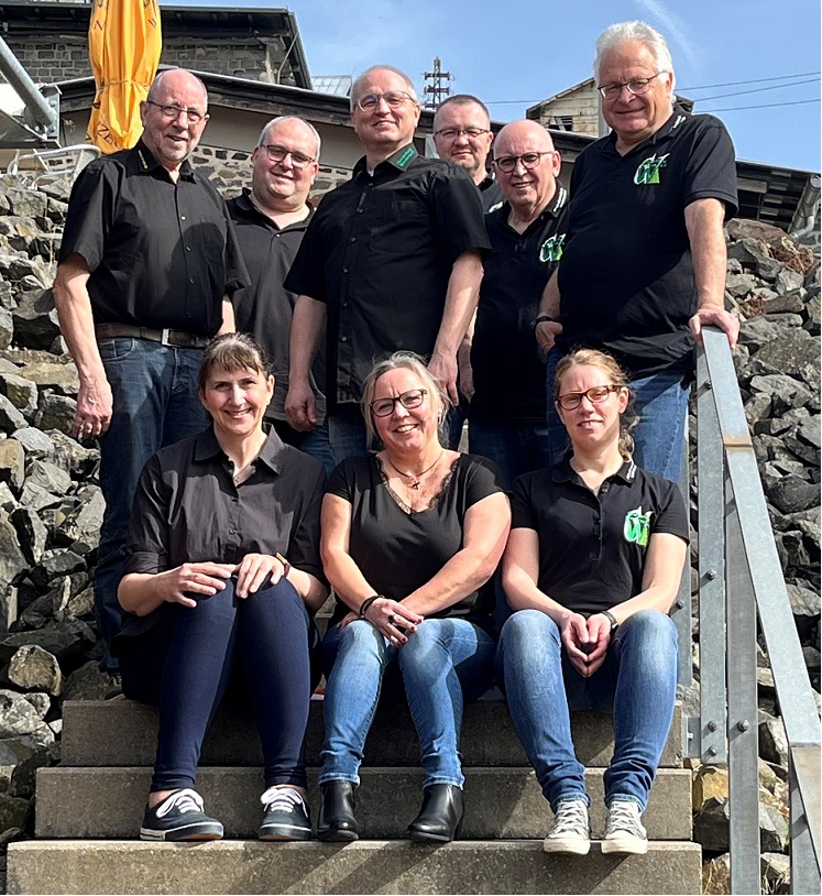 Gruppenfoto des Vorstands des Chorverbands Westerwald auf einer Treppe im Stöffelpark, aufgenommen im April 2024. Acht Personen stehen in zwei Reihen: drei Frauen unten, fünf Männer oben.